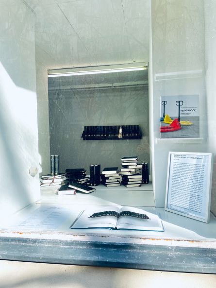 Blick ins Schaufenster, in das die Sonne scheint; Bücherstapel liegen verteilt auf der Fläche hinter dem Fenster. Ganz vorne liegt ein aufgeschlagenes Buch. Hinten an der Wand hängt ein Regal mit einer Reihe in schwarzes Leinen gebundener Gesamtausgabe