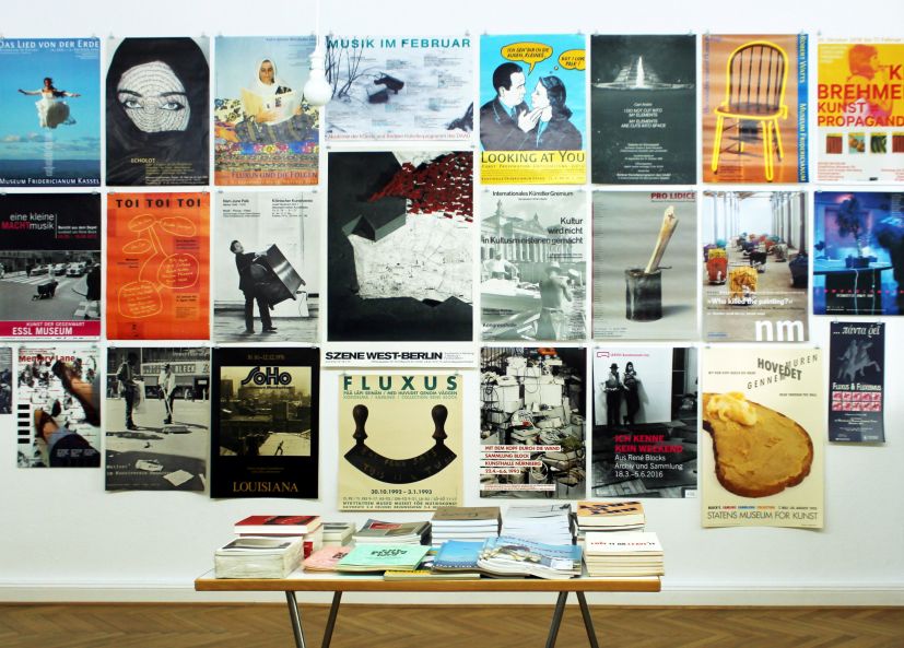 Blick in den Ausstellungsraum: eine Wand, die dicht mit bunten Ausstellungsplakaten behangen ist. Davor steht ein Tisch mit Büchern.