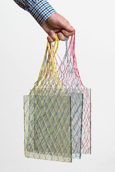 Eine Hand ragt ins Bild und hält 3 Einkaufsnetze in den Grundfarben. In den Netzen ist jeweils eine quadratische Glasplatte
