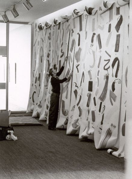 b/w photo shows Eva-Maria Schön installing her work. A long canvas strip with brushstrokes is installed in many loops over a wall