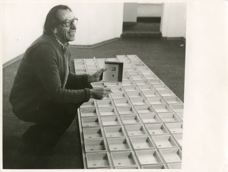 s/w Foto zeigt den Künstler Robert Filliou in der Galerie René Block in Berlin, wie er sein Multiple signiert, das vor ihm auf dem Teppichboden ausgebreitet ist