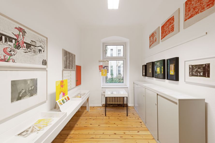 Blick in einen kleinen Raum mit einem niedrigen weißen Display links und einem grauen Schrank rechts. An den Wänden hängen verschiedene Grafiken