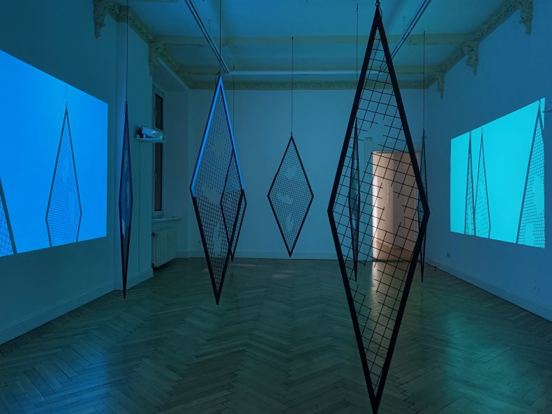 Ausstellungsansicht einer Installation von Sunah Choi. Der Raum ist in bläuliches Licht getaucht. Von der Decke hängen 7 große Rhomben. An die Wände sind Farbflächen projiziert, in denen man die Schatten der Rhomben sieht