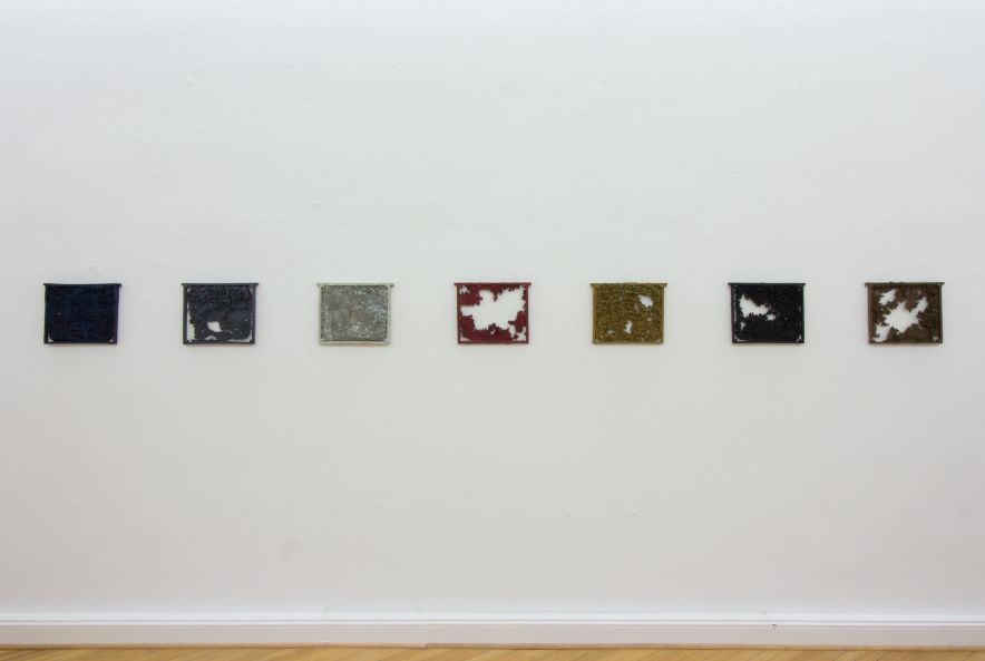 Ausstellungsansicht. 7 Bienenwaben aus Bronze von Björn Nörgaard hängen in einer Reihe an der Wand.