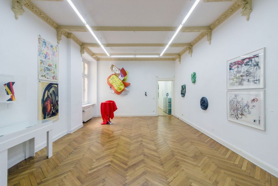 Ausstellungsansicht von Olaf Metzel. Im Raum hängen gerahmte Drucke und Objekte an der Wand, und es steht ein mit rotem Plastik überzogener Stehtisch