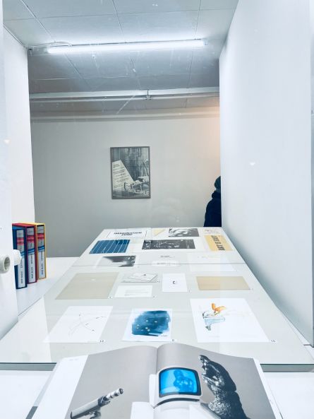 Blick in ein Schaufenster mit einer Neonröhre. Auf einer weißen Fläche hinter dem Fenster liegen verschiedene Zeichnungen, Aquarelle und Drucke. Vorne liegt ein aufgeschlagenes Buch mit einer Abbildung von "Der Denker" von Nam June Paik.