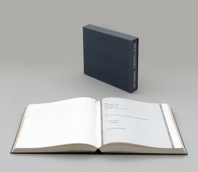 Künstlerbuch von On Kawara. Ein Buch in grauem Leinenschuber steht vor grauem Hintergrund. Auf dem Buchrücken steht in weiß: On Kawara. I AM STILL ALIVE. Ein weiteres Exemplar liegt aufgeschlagen im Vordergrund.