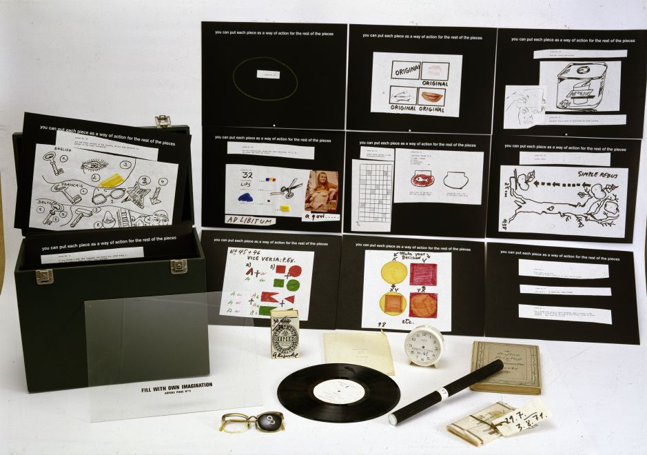 Edition by Arthur Köpcke. A small black case with black index cards on which texts and drawings are glued. 9 of the index cards are spread out next to the suitcase. Several objects are also lying next to it.