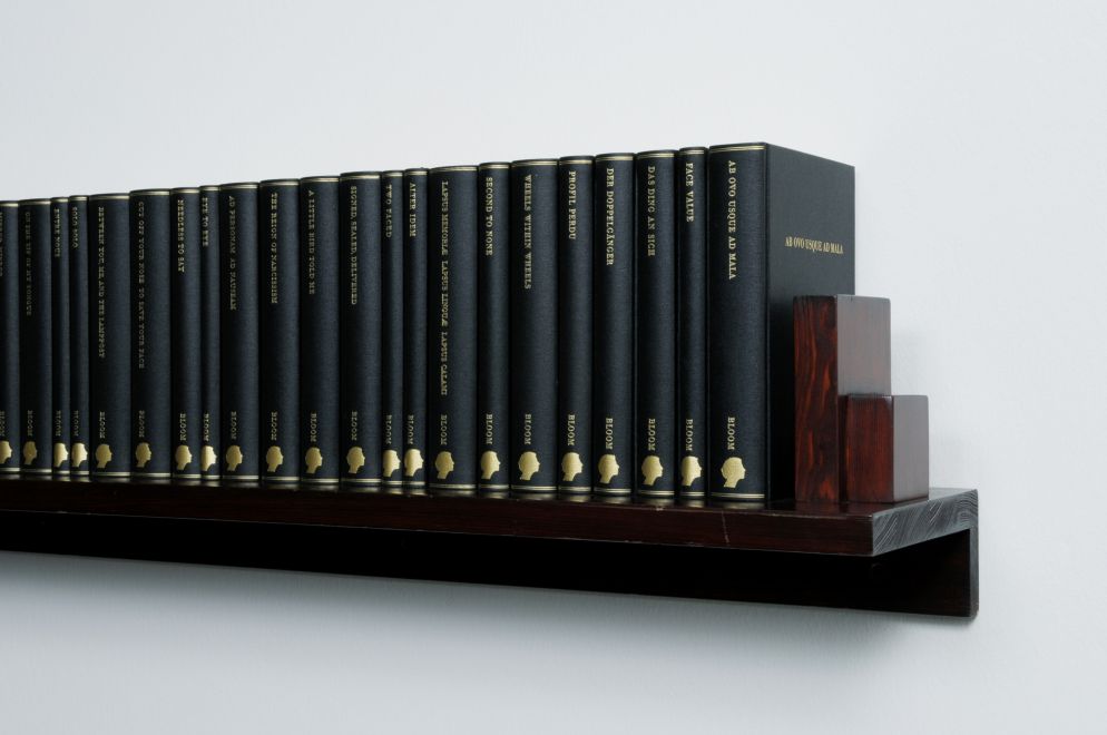 Edition der Künstlerin Barbara Bloom. Ein Wandregal aus dunkelbraunem Holz hängt an der Wand. Darauf stehen 38 in schwarzes Leinen gebundene Bücher mit Goldprägung auf den Buchrücken. Detailansicht, die einen Teil des Regals zeigt.