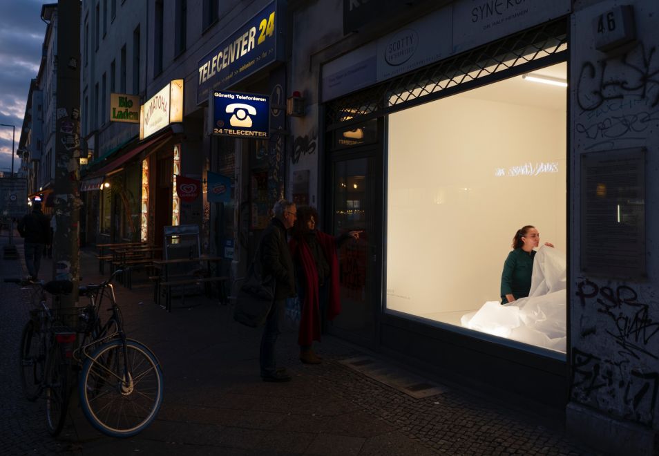 Artist Mehtap Baydu sitting inside a Berlin shop window during her performance ATEM, inflating a large translucent balloon over 18 days using only her breath. Two people in front of the window are watching her.