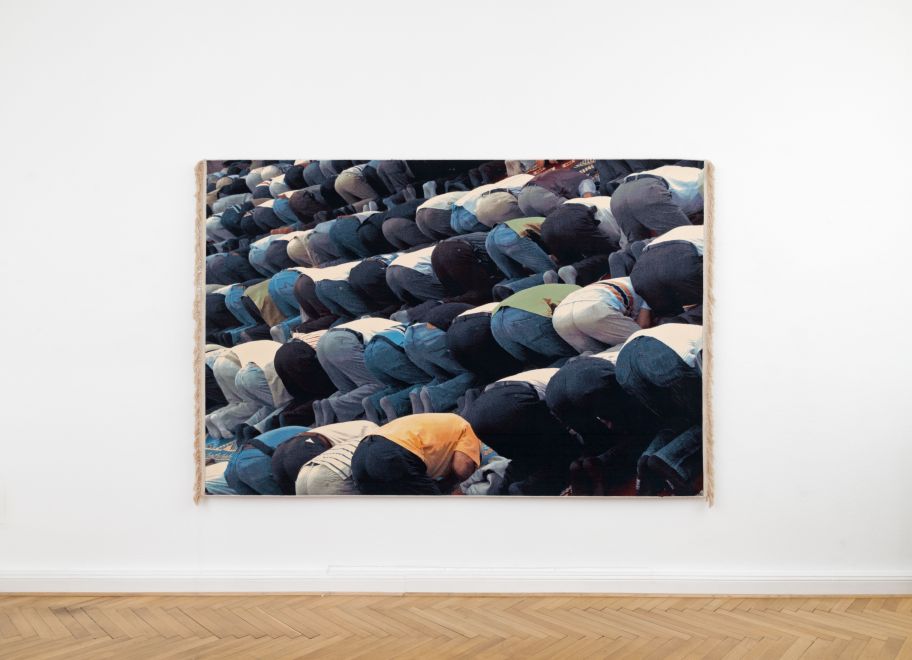 Ein großer Wandteppich von Mehtap Baydu an einer weißen Galeriewand zeigt Reihen von Männern, die sich im Freitagsgebet nach vorne beugen, von hinten gesehen in verschiedenfarbiger Kleidung.