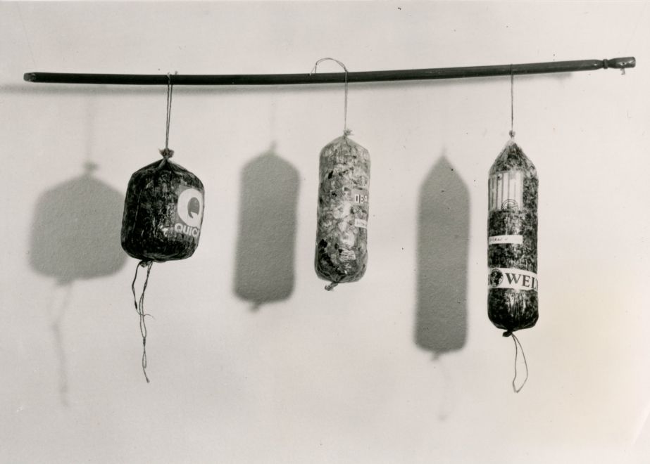 Multiple von Dieter Roth. s/w Foto: 3 Würste in verschiedenen Größen hängen an einer Stange. Sie sind mit zerkleinerten Zeitungen gefüllt. Auf den Würsten sind Logos geklebt: Der Spiegel, Bild, Welt, usw.