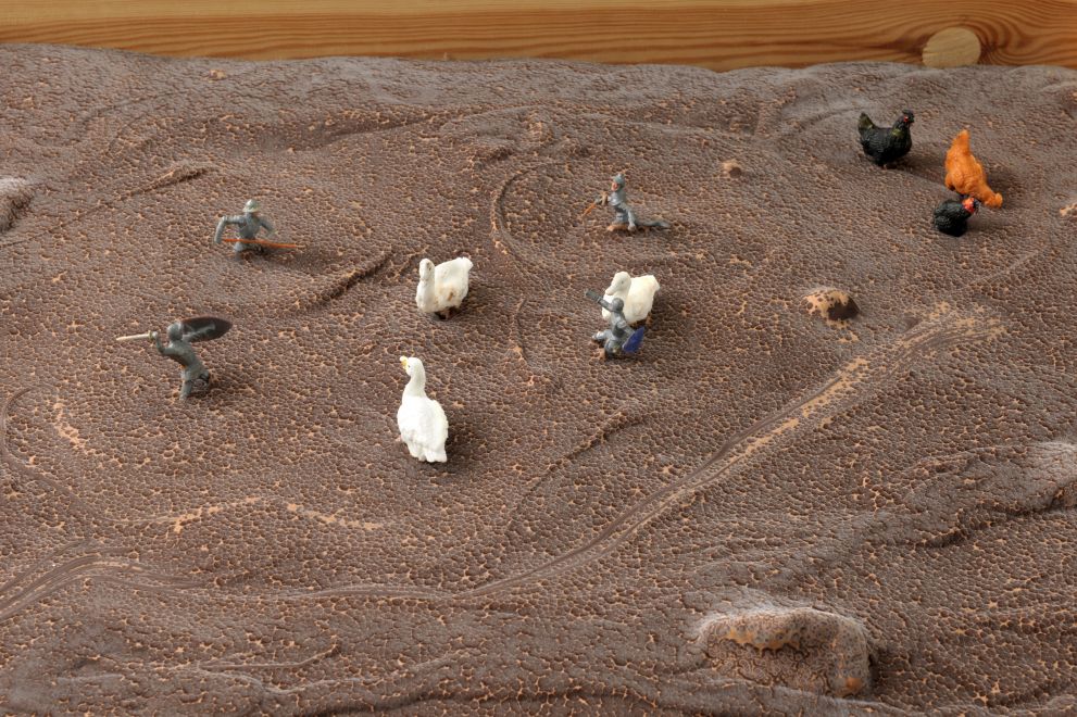 Multiple von Dieter Roth. In eine flache Holzkiste wurde eine unebene Fläche mit Schokolade gegossen. Darin sind kleine Ritterfiguren mit Schwertern und gleich große Enten- und Hühnerfiguren platziert