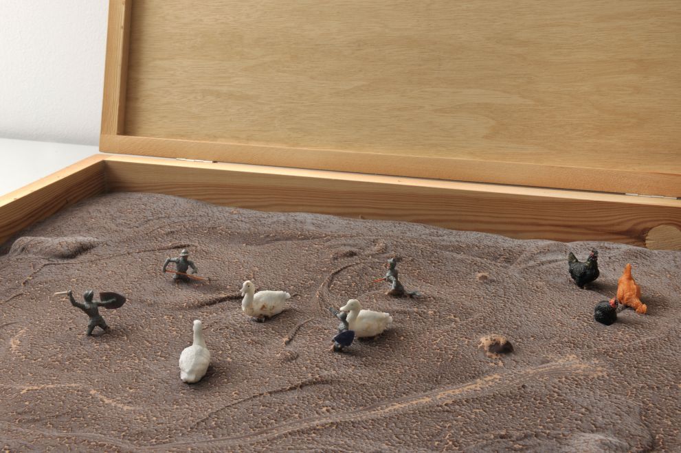Multiple von Dieter Roth. In eine flache Holzkiste wurde eine unebene Fläche mit Schokolade gegossen. Darin sind kleine Ritterfiguren mit Schwertern und gleich große Enten- und Hühnerfiguren platziert
