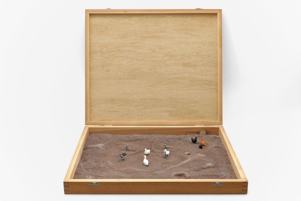 Multiple von Dieter Roth. In eine aufgeklappte flache Holzkiste wurde eine unebene Fläche mit Schokolade gegossen. Darin sind kleine Ritterfiguren mit Schwertern und gleich große Enten- und Hühnerfiguren platziert