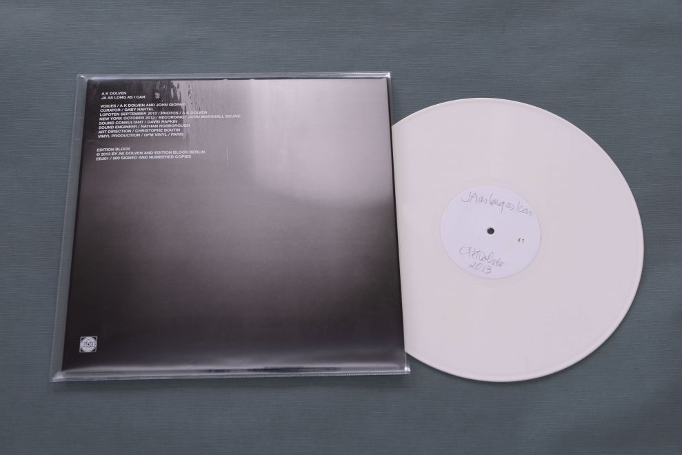 LP der Künstlerin A K Dolven. Auf der Hülle ist ein verschwommenes schwarz-weiß Foto zu sehen, darüber ein weißer Text in der linken oberen Ecke. Die weiße LP ist halb aus der Hülle herausgezogen. Auf dem Label steht handschriftlich: JA as long as I can. A K Dolven. 2013