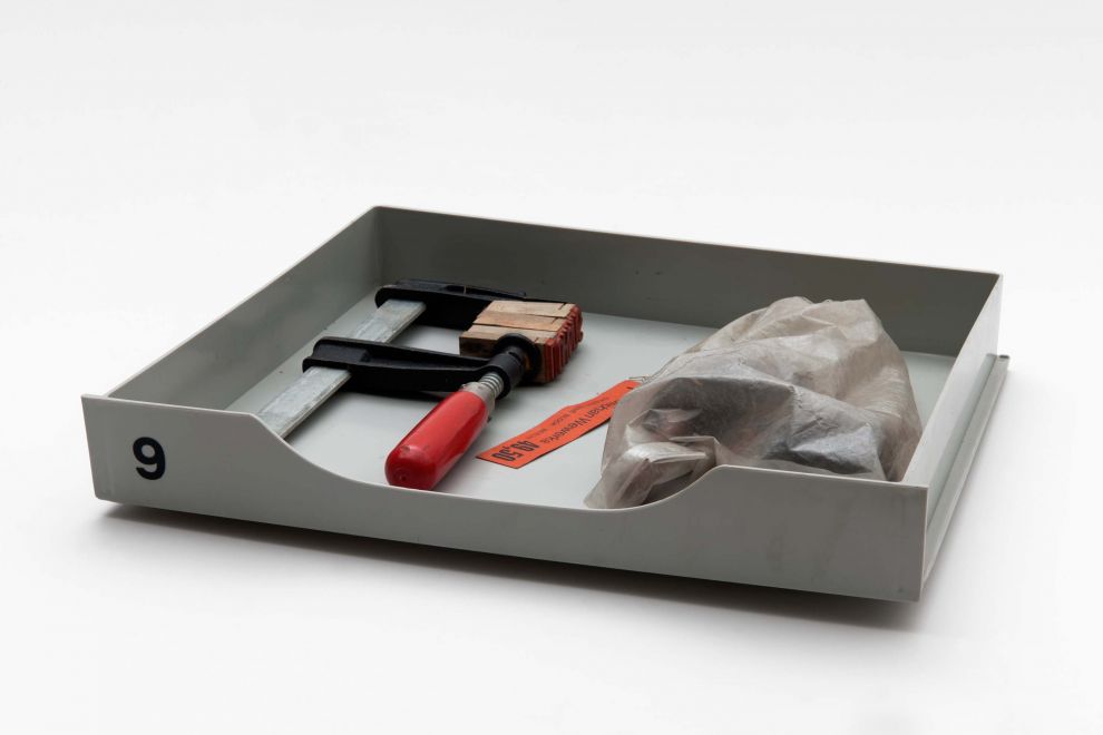 Edition von Bernd Lohaus. In einer grauen Plastikschublade liegt eine Schraubzwinge mit eingeklemmten Stempelbuchstaben. Daneben liegt eine Edition von Wewerka in einer Plastiktüte