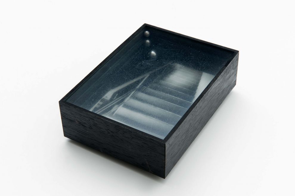 Multiple von Gerhard Richter. Kleiner schwarzer Holzkasten, auf dessen Boden ein s/w Foto seiner Ateliertreppe. In der Kiste sind 3 Glasscheiben auf Ebenen übereinander montiert. Auf jeder Ebene ist eine kleine Stahlkugel.