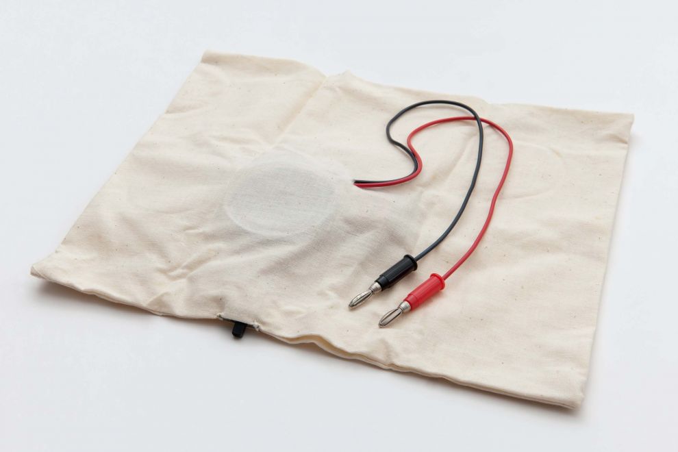 Edition von Hansjoachim Dietrich. Ein Kissenbezug aus hellem Leinen, in den ein schwarzer Kippschalter genäht ist. Aus dem Kissen kommen ein rotes und schwarzes Kabel mit Lamellensteckern