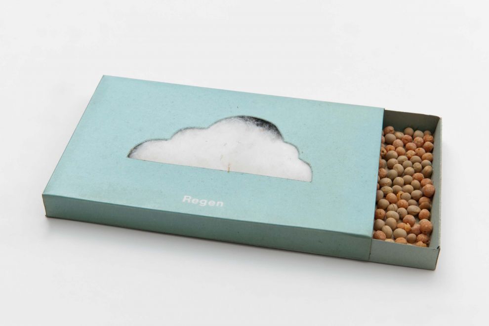 Edition by KP Brehmer. A light blue, half-opened box containing dried peas. The shape of a cloud is punched out on the top, through which white absorbent cotton can be seen. Regen (rain) is written in white under the cloud.