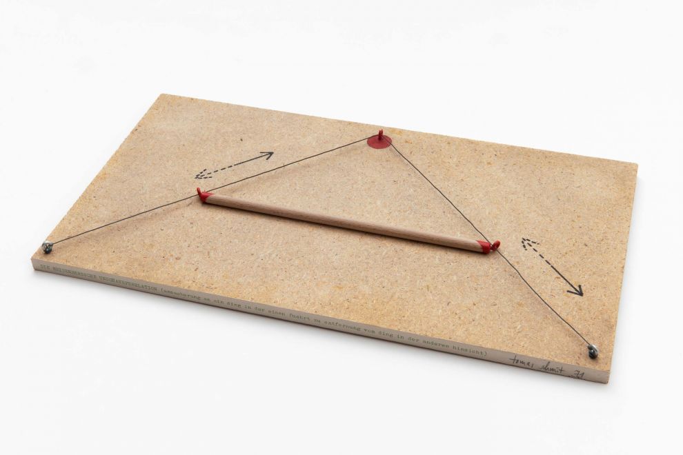 Multiple by Tomas Schmit. A string is stretched at a right angle on a chipboard panel, with a movable wooden stick stretched between them. Arrows are painted along the string. On the front edge of the board is written: THE HEISENBERG'S UNSCHÄRFEREALTION (approaching a thing in one way leads to distance from the thing in another)
