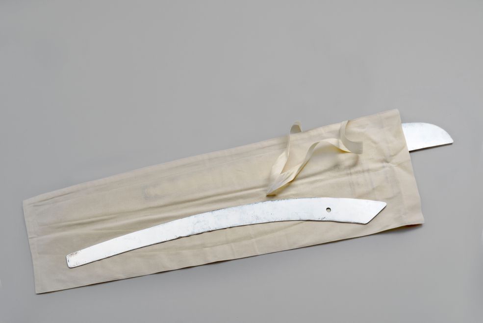 Edition von Ayse Erkmen. Eine Schneiderschablone aus Silber in Form einer Säbelklinge liebt auf einer hellen Hülle aus Leinen. Eine zweite Schablone kuckt aus der Hülle hervor.