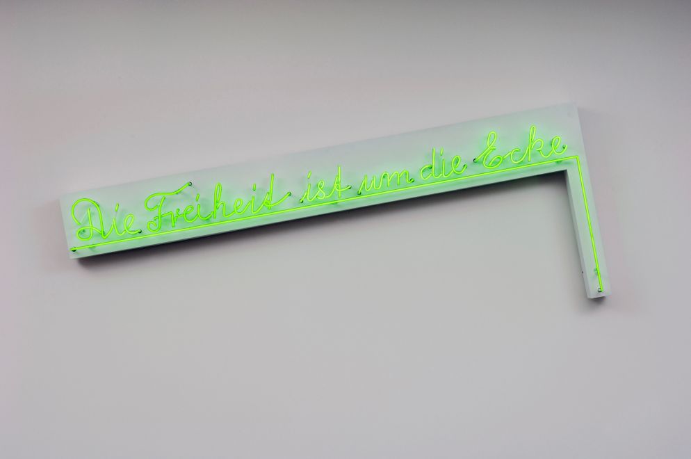 Wall object by Henning Christiansen. Handwritten in green neon letters, DIE FREIHEIT IST UM DIE ECKE is mounted on a metal body. The sentence is underlined by a neon line that bends downwards at the end - around the corner