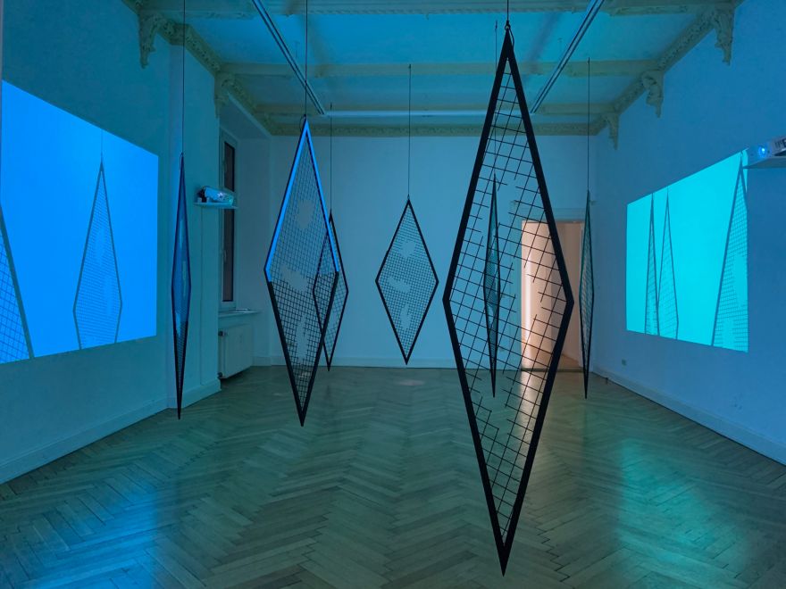 Installation Karo von Sunah Choi. In einem Galerieraum hängen 7 rautenförmige Gitterelemente von der Decke. Auf beiden Seiten befinden sich Projektoren, die jeweils eine Farbfläche auf die gegenüberliegende Wand projizieren, in der man Schatten der Gitterelemente sieht. Der abgedunkelte Raum ist dadurch in bläuliches Licht getaucht.