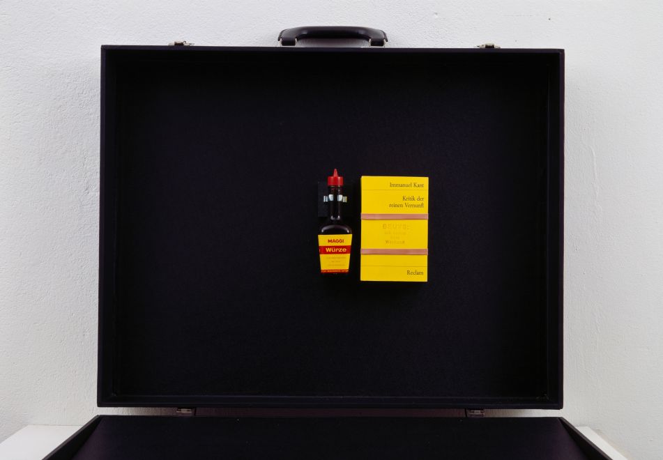 Multiple by the artist Joseph Beuys, consisting of a Maggi bottle and the Reclam book Immanuel Kant, Kritik der reinen Vernunft, stamped in red ink with BEUYS: ich kenne kein weekend. The bottle and book are mounted on the lid of a black suitcase.