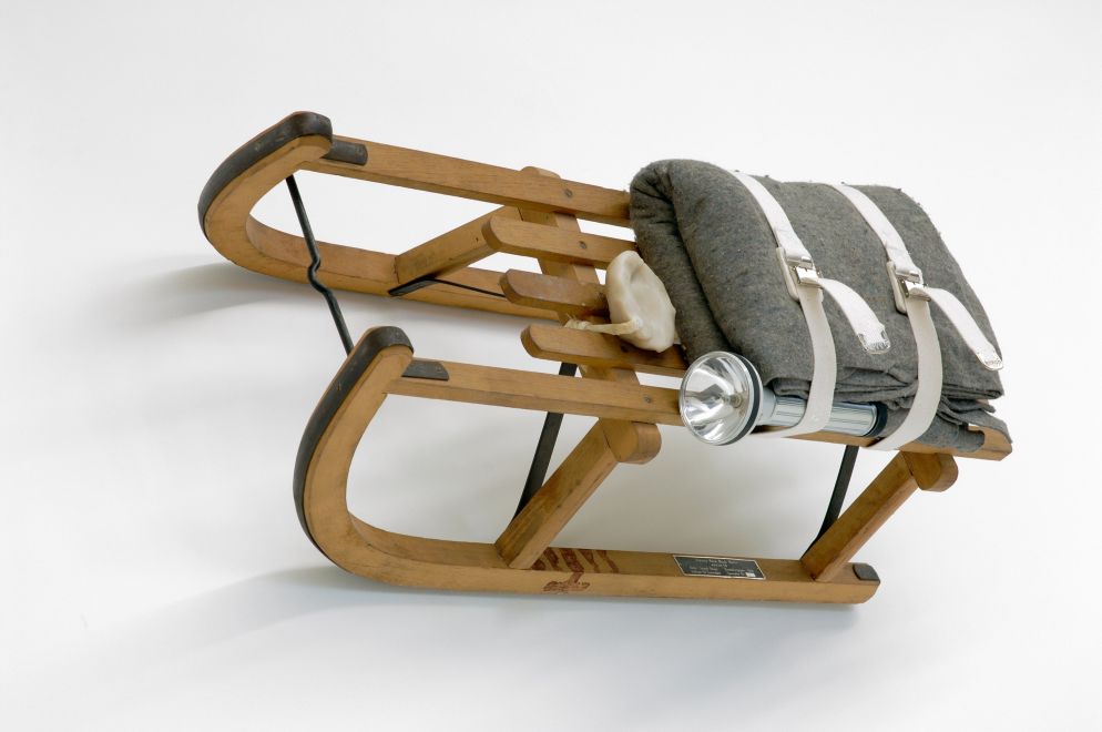 Multiple von Joseph Beuys. Ein Holzschlitten, auf dem eine kleine Fettplastik liegt und eine Filzdecke und Taschenlampe mit 2 Bändern befestigt sind.
