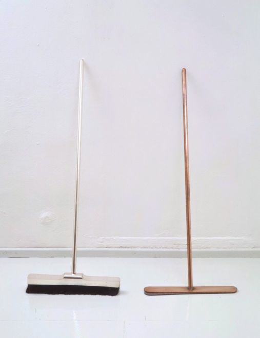 Kunstedition des Künstlers Joseph Beuys. In einem weißen Galerieraum lehnen zwei Besen nebeneinander an einer Wand. Der Besen auf der linken Seite ist silbern mit schwarzem Pferdehaar, der Besen auf der rechten Seite ist kupferfarben mit einem Stück Filz anstelle der Haare.