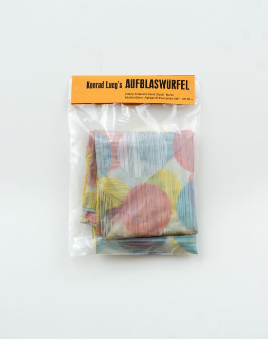 Edition von Konrad Lueg. Ein Plastikbeutel mit einem zusammengefalteten Plastikwürfel mit weißen, rosa, gelben und hellblauen Punkten. Auf dem Label des Beutels steht KONRAD LUEGS AUFBLASWÜRFEL Edition 6 Galerie René Block Berlin