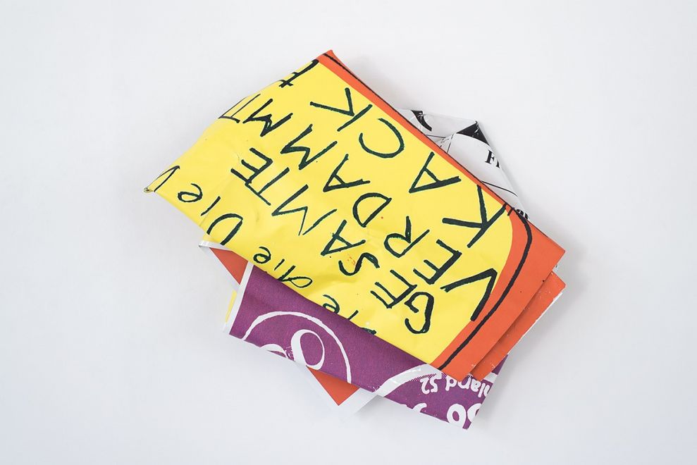 Edition by Olaf Metzel. Object hangs on a wall. Aluminum plate printed on both sides, irregularly folded into a smaller object so that a handwritten quote by Dieter Roth can be read: Die GESAMTE VERDAMMTE KACKE