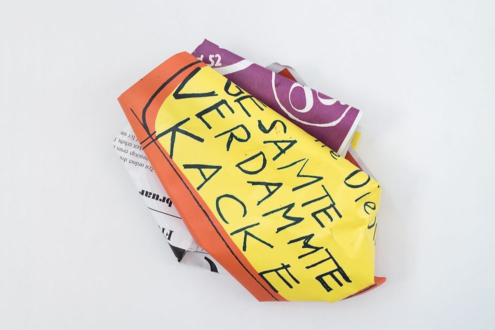 Edition by Olaf Metzel. Object hangs on a wall. Aluminum plate printed on both sides, irregularly folded into a smaller object so that a handwritten quote by Dieter Roth can be read: Die GESAMTE VERDAMMTE KACKE