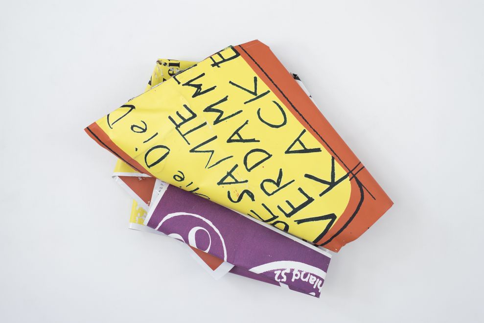 Edition by Olaf Metzel. Object hangs on a wall. Aluminum plate printed on both sides, irregularly folded into a smaller object so that a handwritten quote by Dieter Roth can be read: Die GESAMTE VERDAMMTE KACKE