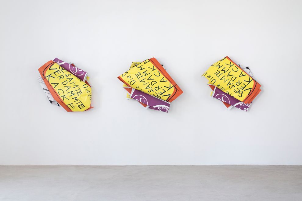 Edition by Olaf Metzel. 3 objects hanging in a gallery space. Aluminum plates printed on both sides, irregularly folded into smaller objects, so that a handwritten quote by Dieter Roth can be read: Die GESAMTE VERDAMMTE KACKE (The whole damn shit)