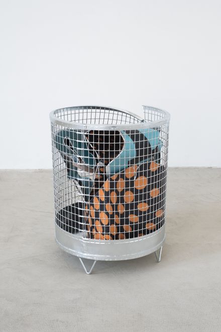 Edition von Olaf Metzel. In einem runden Papierkorb aus verzinktem Stahlgitter ist eine beidseitig bedruckte Aluminiumplatte, die so gefaltet ist, dass sie aussieht wie ein zerknülltes Blatt Papier.