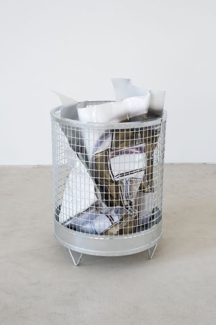 Edition von Olaf Metzel. In einem runden Papierkorb aus verzinktem Stahlgitter ist eine beidseitig bedruckte Aluminiumplatte, die so gefaltet ist, dass sie aussieht wie ein zerknülltes Blatt Papier.