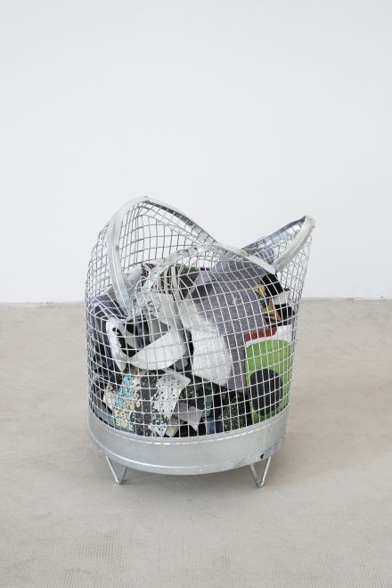 Edition von Olaf Metzel. In einem runden Papierkorb aus verzinktem Stahlgitter ist eine beidseitig bedruckte Aluminiumplatte, die so gefaltet ist, dass sie aussieht wie ein zerknülltes Blatt Papier. Der Papierkorb ist seinerseits eingedrückt