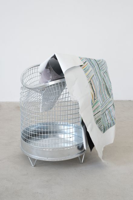 Edition von Olaf Metzel. In einem runden Papierkorb aus verzinktem Stahlgitter ist eine beidseitig bedruckte Aluminiumplatte, die so gefaltet ist, dass sie aussieht wie ein zerknülltes Blatt Papier, das über dem Rand hängt