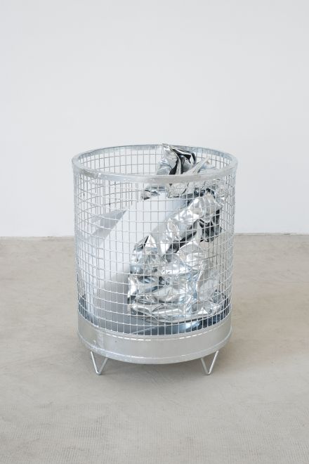 Edition von Olaf Metzel. In einem runden Papierkorb aus verzinktem Stahlgitter ist eine bedruckte Aluminiumplatte, die so gefaltet ist, dass sie aussieht wie ein zerknülltes Blatt Papier