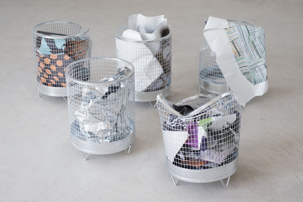 Edition von Olaf Metzel. In 5 runden Papierkörben aus verzinktem Stahlgitter sind beidseitig bedruckte Aluminiumplatten, die so gefaltet ist, dass sie aussehen wie zerknülltes Papier. Manche der Körbe sind eingedrückt