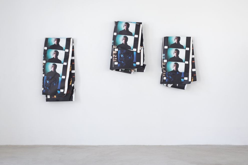 Edition von Olaf Metzel. 3 Wandskulpturen hängen in einem Galerieraum. Es sind beidseitig bedruckte Aluminuimplatten, die unregelmäßig zu kleineren Objekten gefaltet sind, so dass Filmstreifen mit Fotos von Andy Warhol zu sehen sind