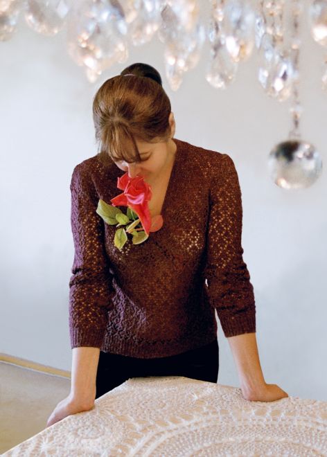 Offsetdruck von Aydan Murtezaoglu. Eine Frau steht an einem Tisch mit Häckeldecke, sie hat die Hände auf den Tisch gestützt, blickt nach unten und hat eine rote Rose im Mund. Über ihrem Kopf noch Teile eines Kristall Kronleuchters