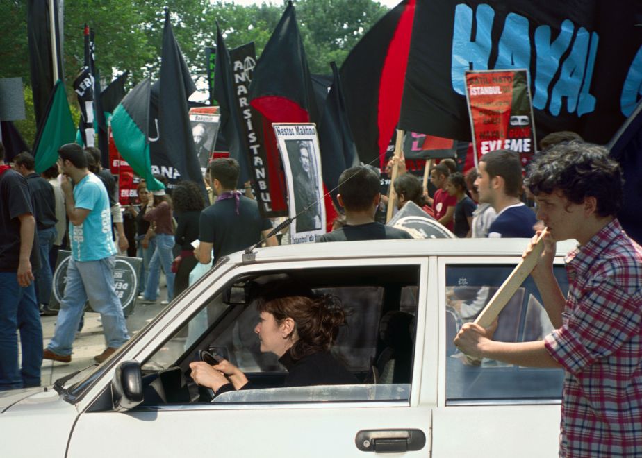Offsetdruck von Aydan Murtezaoglu. Eine Frau am Steuer eines weißen Autos fährt mitten durch eine Demonstration