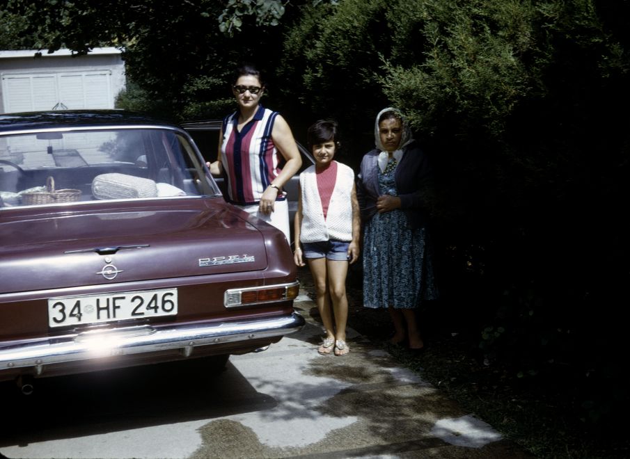 Offsetdruck von Aydan Murtezaoglu. Eine Aufnahme aus den 1970ern zeigt einen alten roten Opel von hinten, neben ihm stehen 2 Frauen und ein Mädchen.