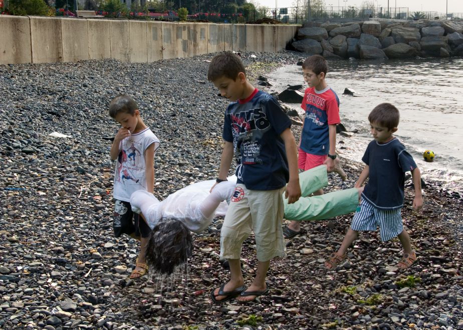 Offsetdruck von Aydan Murtezaoglu. Fotomontage. 4 Kinder tragen eine nass-triefende erwachsene Person mit dem Gesicht nach unten an Händen und Beinen scheinbar aus einem See.