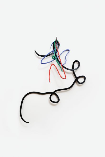 Edition von Navid Nuur. Geschwungene Metallformen in den Farben Grün, Rot, Blau und Schwarz hängen an einem silbernen Mehrfarbkugelschreiber an einer weißen Wand. Die Formen sehen aus wie bunte Kritzeleien