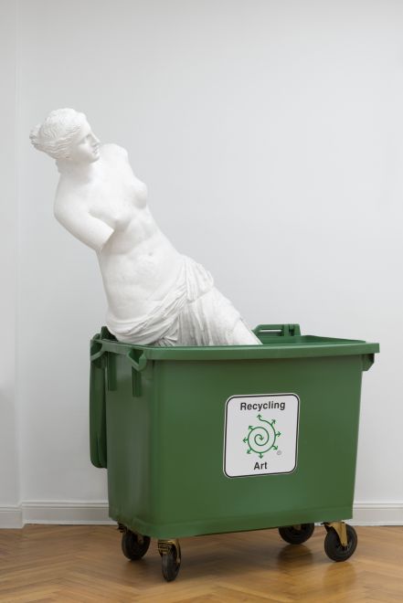 Edition des Künstlers Bjørn Nørgaard in einem Galerieraum. Aus einer grünen Recycling-Tonne ragt der weiße Gipsabguss der berühmten Venus von Milo. Ein Aufkleber auf der Tonne zeigt eine Spirale, von der mehrere Pfeile ausgehen