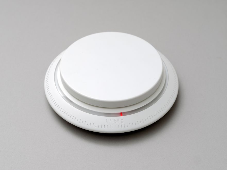 Video-Objekt der Künstlerin Ebru Özsecen. Eine weiße runde Küchenwaage, die als Verpackung für die DVD dient. Als Skalierung ist 0/158 g angegeben.